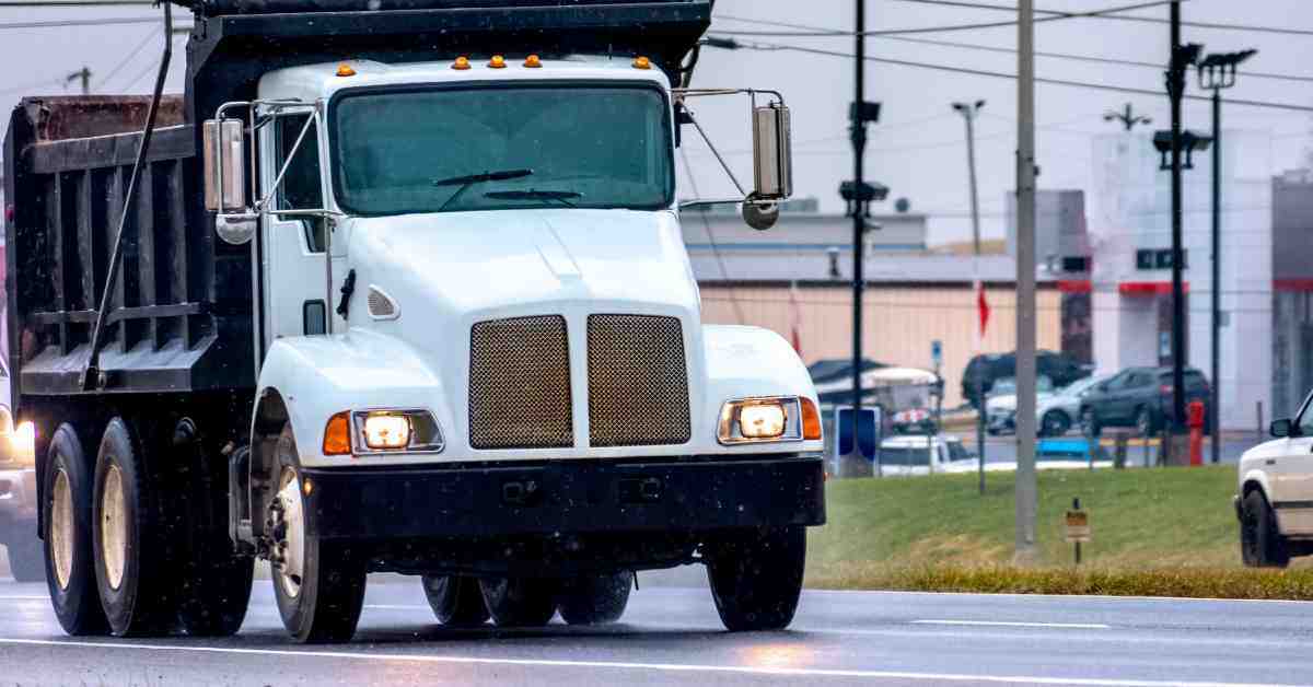 A dump truck drives down the road on a wet day. The front of the vehicle is white, while the rest is black.