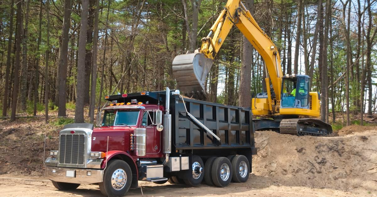 A yellow construction excavator drops sand into the back of a dump truck. Both vehicles are close to a forest.