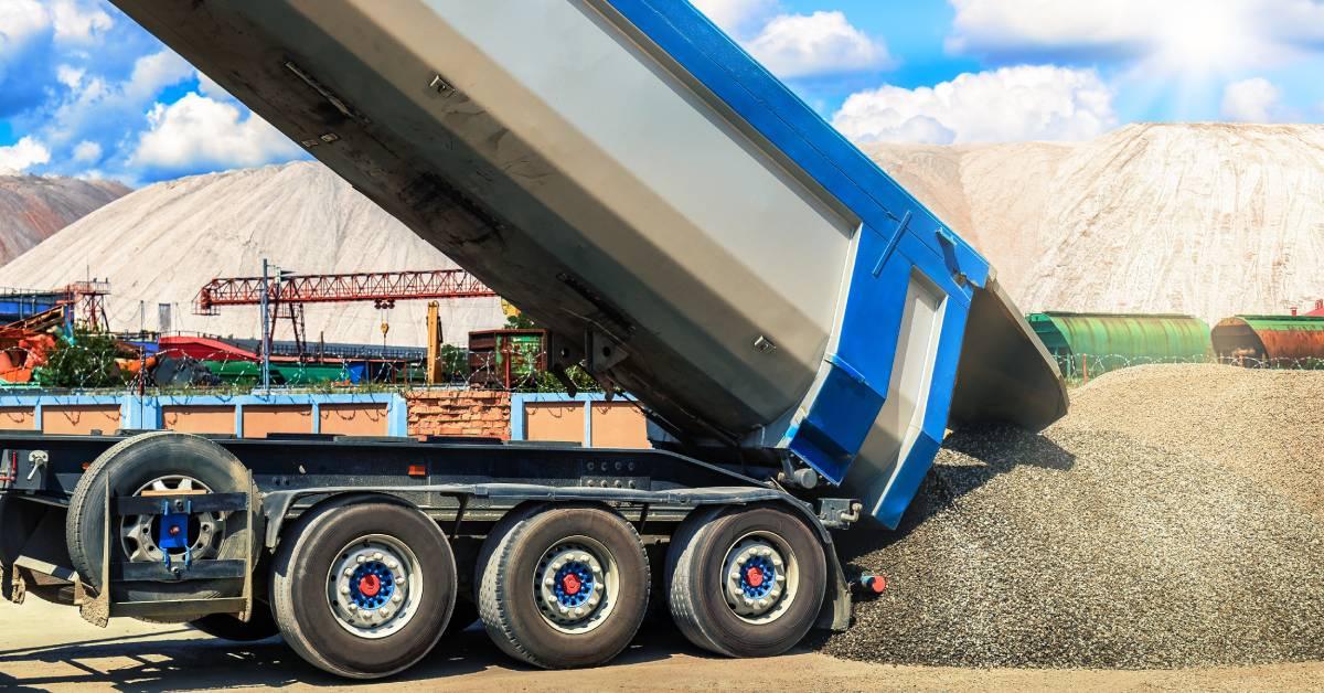 A blue and white dump truck dumps a load of gravel onto the ground. A blue sky with clouds is in the background.