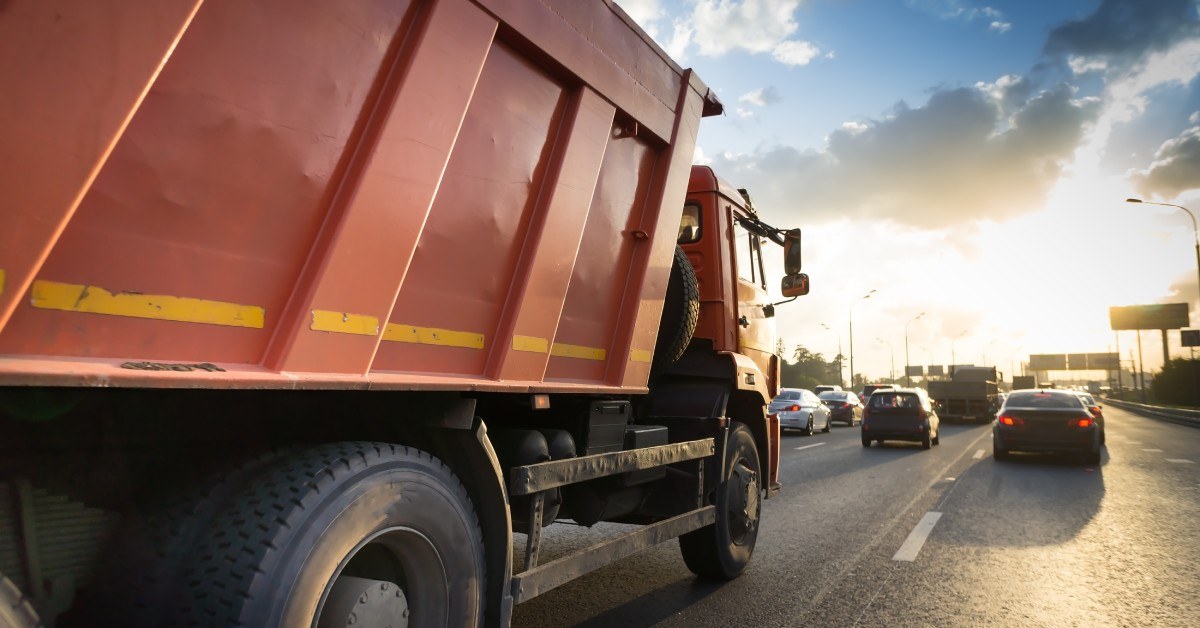 A red dump truck drives down the highway behind other cars as the sun starts to go down. Highway signs stand in the distance.