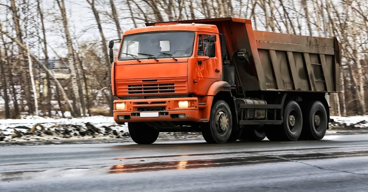 A red dump truck drives on a road during the winter. Snow is on the side of the road, while the trees are bare.