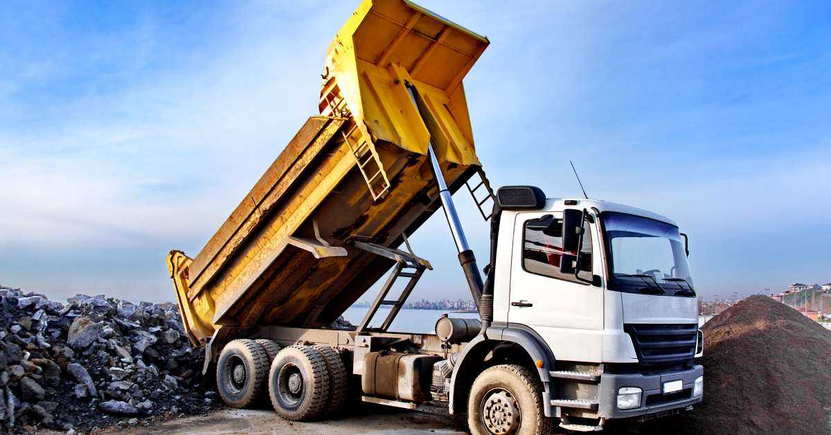 A dump truck empties a load into a pile of rocks on a job site. The driver has parked the truck next to a pile of dirt.