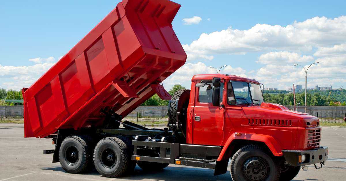 A red dump truck with its back load elevated sits in an empty parking lot. A blue sky is in the background.