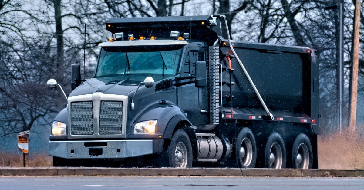 A black dump truck drives on a road with its headlights on. Several trees stand in the background behind it.