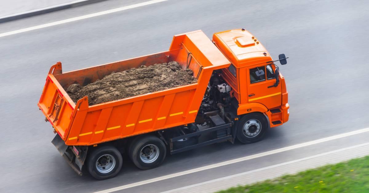 An orange dump truck drives on a highway, carrying dirt in the back. Grass is on the side of the road.