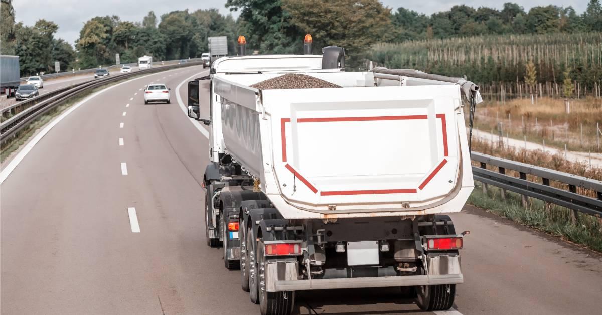 The back of a white dump truck traveling on a highway and heading toward a curve in the road. It is carrying gravel.