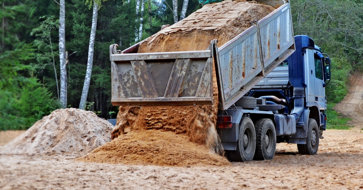 A large blue-colored dump truck dumping a bed full of dirt into a spacious construction site. Trees stand in the background.