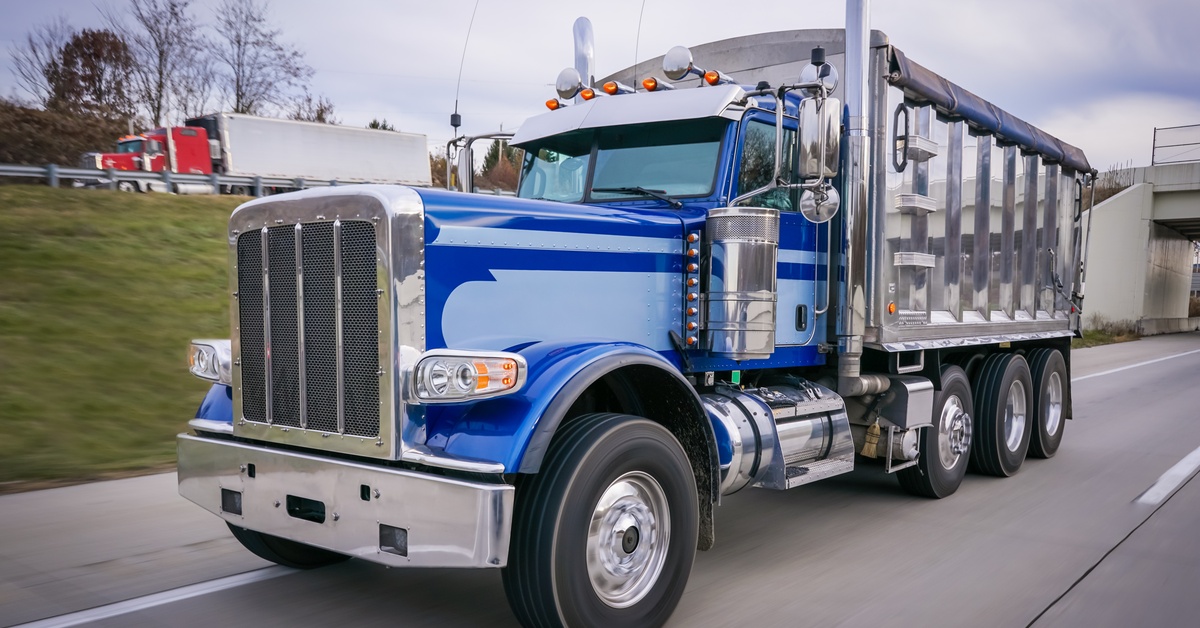 A blue dump truck drives along a highway with a silver dump body as a red semi passes in the background.