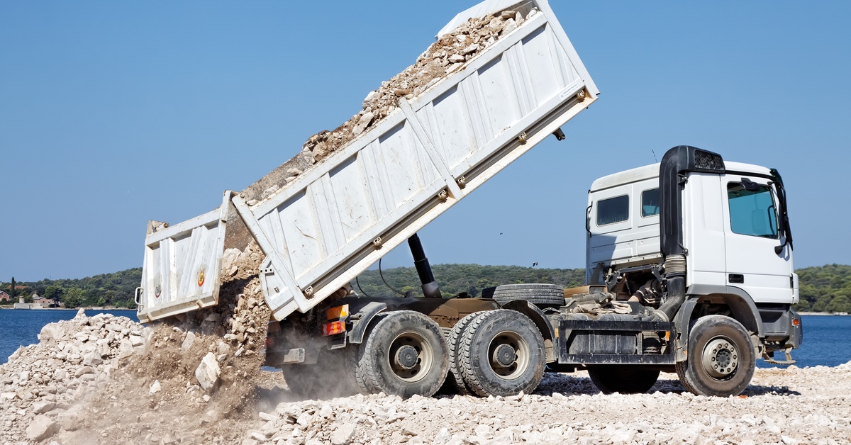 A large white-colored dump truck parked at a construction site near a lake dumps a load of rocks and gravel.