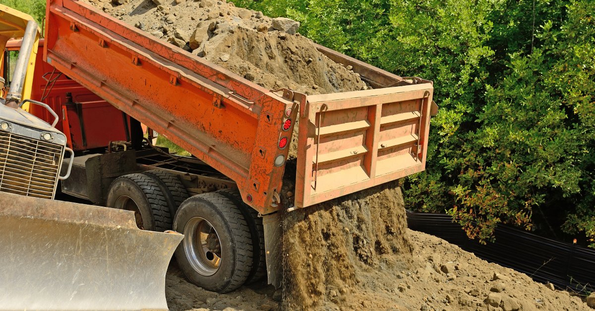 An orange-colored dump truck dumps a load of dirt next to another machine at a construction site surrounded by trees.