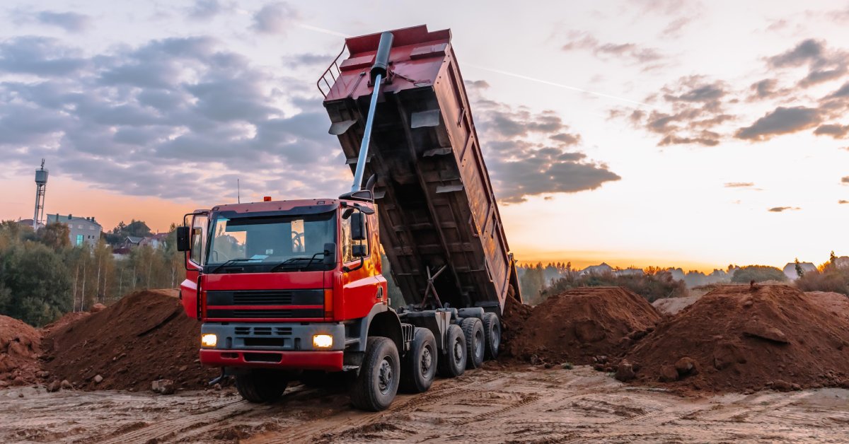 A red dump truck is parked and unloading a bed full of dirt near other dirt piles at an excavation site.