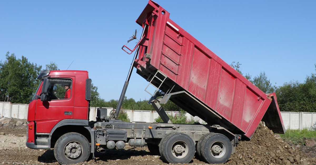 A large red-colored dump truck sits parked in the middle of a construction site while dropping a load of dirt.