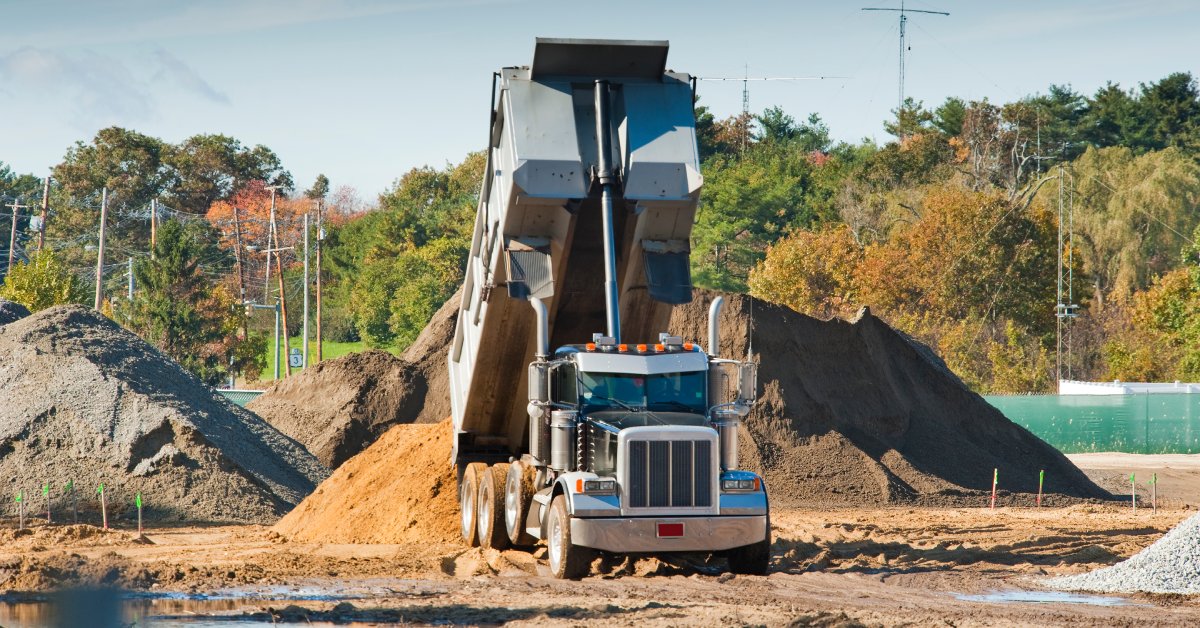 A large black-and-silver dump truck dumps a bed full of dirt into a pile in the middle of a construction site.