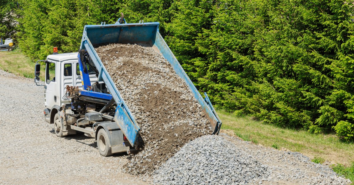 A white and blue-colored dump truck unloads its bed full of gravel into a pile at a construction site.
