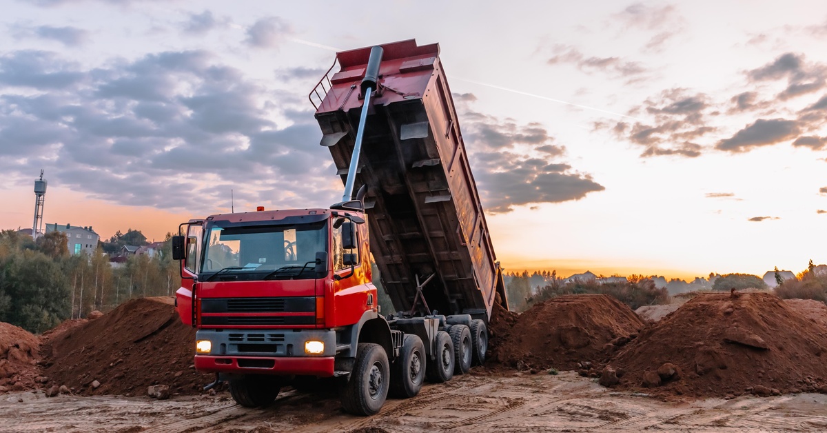 A large red-colored dump truck is parked at an excavation site while dumping loads of dirt and gravel into a pile.