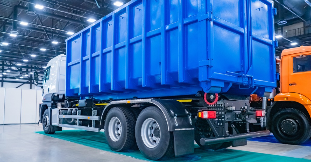 A blue dump truck with a large steel bed is parked inside a warehouse, with bright lights overhead and another truck nearby.