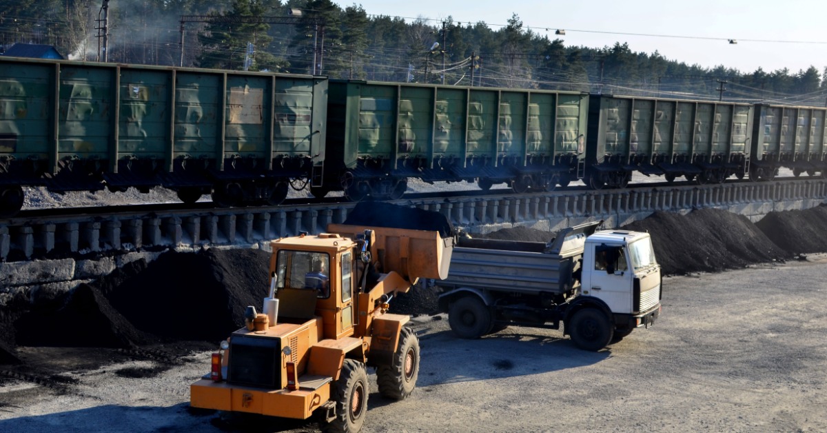 A wheel loader fills a dump truck with coal beside railcars and stockpiles at an industrial loading yard.