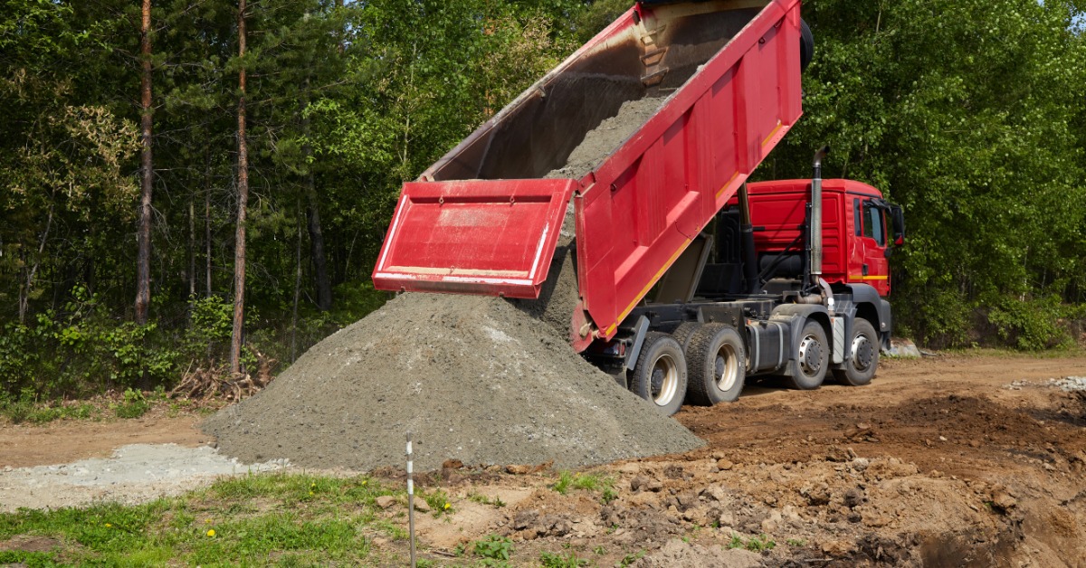 A red dump truck unloads a large pile of light gray gravel on a dirt site, surrounded by green trees and grass.