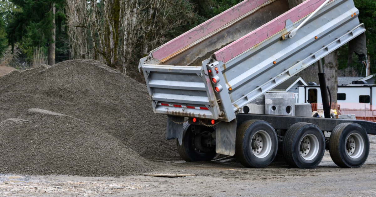 A dump truck raises the truck bed and unloads a pile of gravel at a construction site with trees in the background.