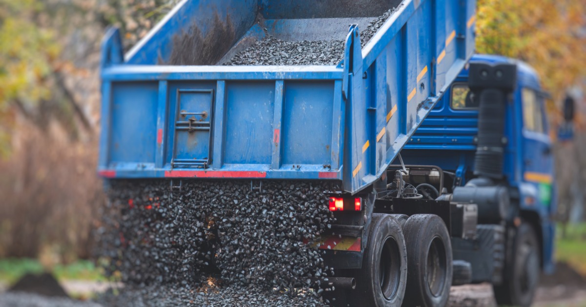 A blue dump truck unloads dark gravel from a raised bed onto a roadside surface with trees in the background.
