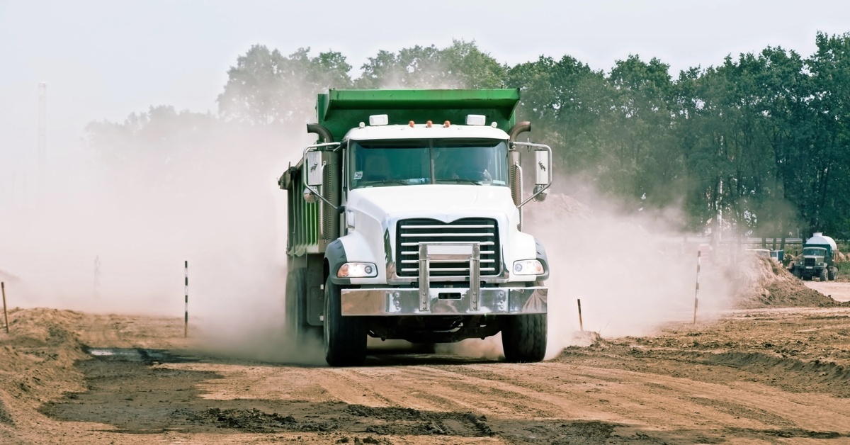 A white and green dump truck drives down a dusty dirt road near a wooded construction site on a cloudy day.