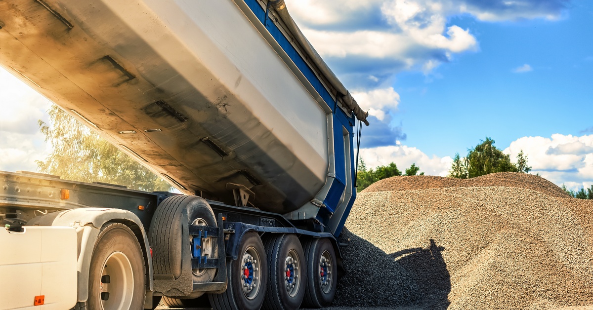 A raised dump trailer unloads gravel beside a large aggregate pile under a bright blue sky at a work site.