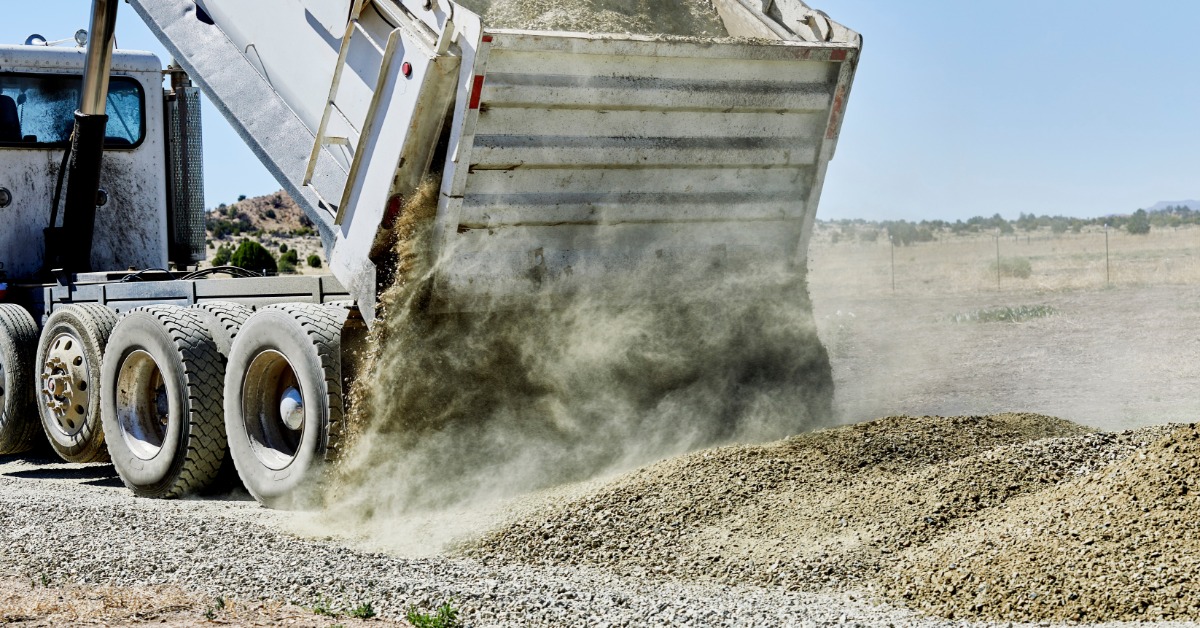 A dump truck releases gravel as a thick dust cloud rises behind a growing pile on a dry roadside worksite.