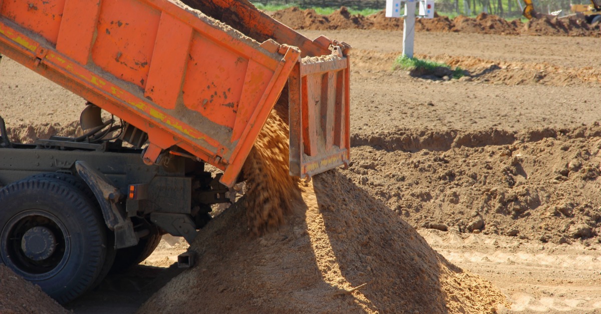 An orange dump truck unloads sand into a pile at a sunlit construction site with an excavator in the background.